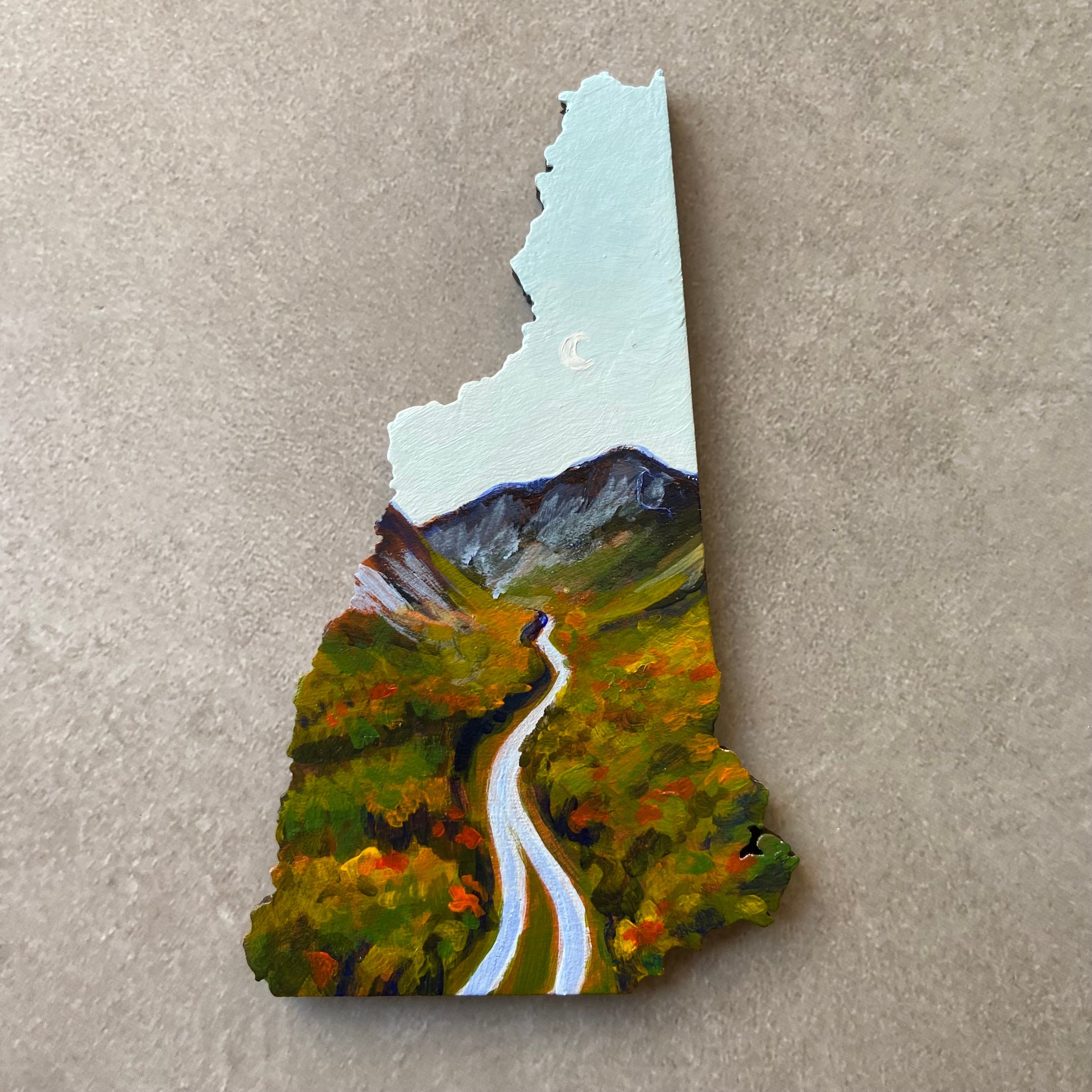 A New Hampshire-shaped painting of a highway extending into the distance towards mountains under a bright blue sky. It is the beginning of fall foliage season, with red and orange scattered in the forest, and there is a subtle crescent moon in the sky. 