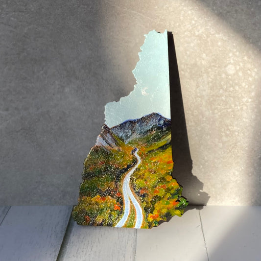 A New Hampshire-shaped painting of a highway extending into the distance towards mountains under a bright blue sky. It is the beginning of fall foliage season, with red and orange scattered in the forest, and there is a subtle crescent moon in the sky.  The painting is upright in a patch of sun.
