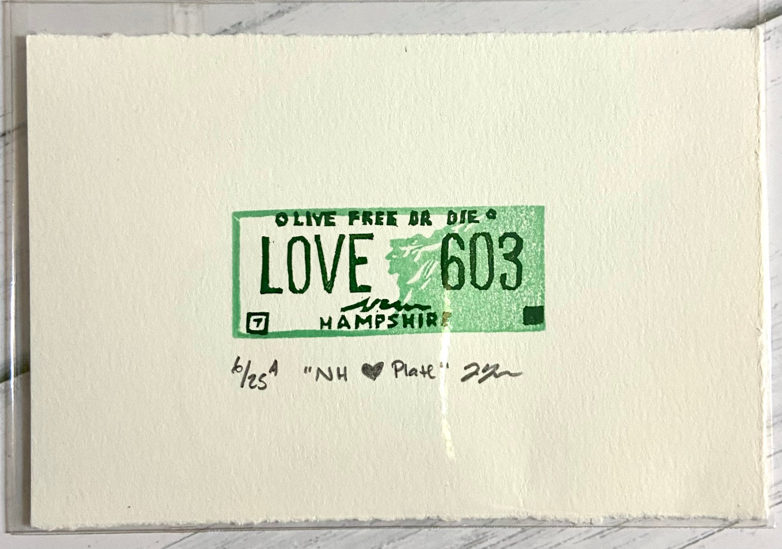 A miniature block print eraser print with the text 'LOVE 603' on a piece of paper, featuring green ink on a white background with a signature at the bottom.