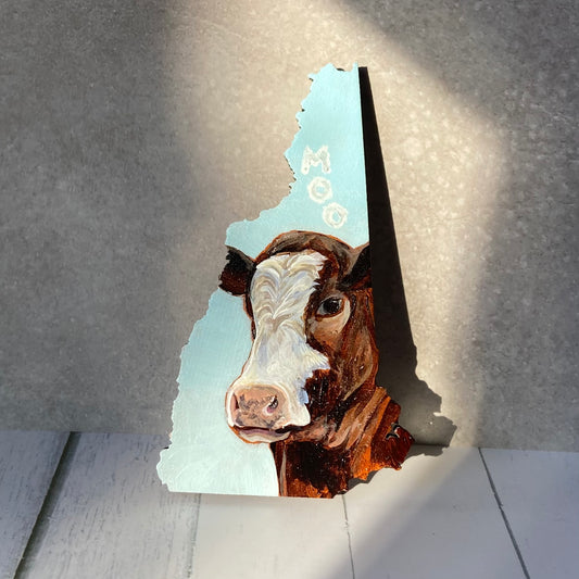 A New Hampshire-shaped painting depicting a brown and white cow's face looking at the viewer. The sky-blue background has three letter-shaped clouds over the cow's head, reading "MOO". The painting is standing upright in a sunny spot.