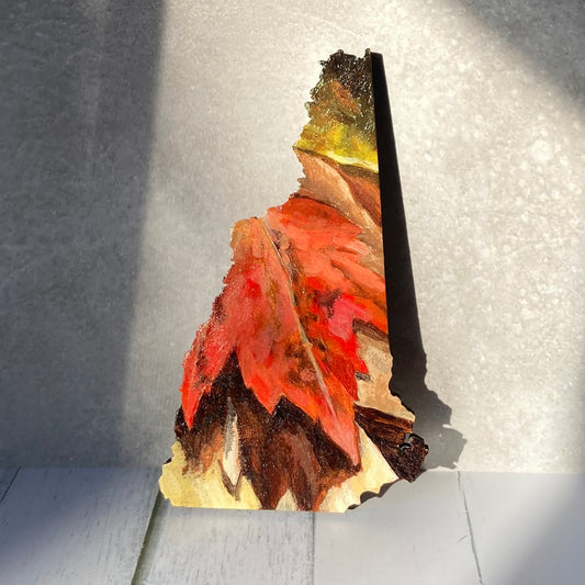A New Hampshire-shaped painting depicting a red maple leaf on the ground among other fall leaves. The painting is propped up in a sunny spot.