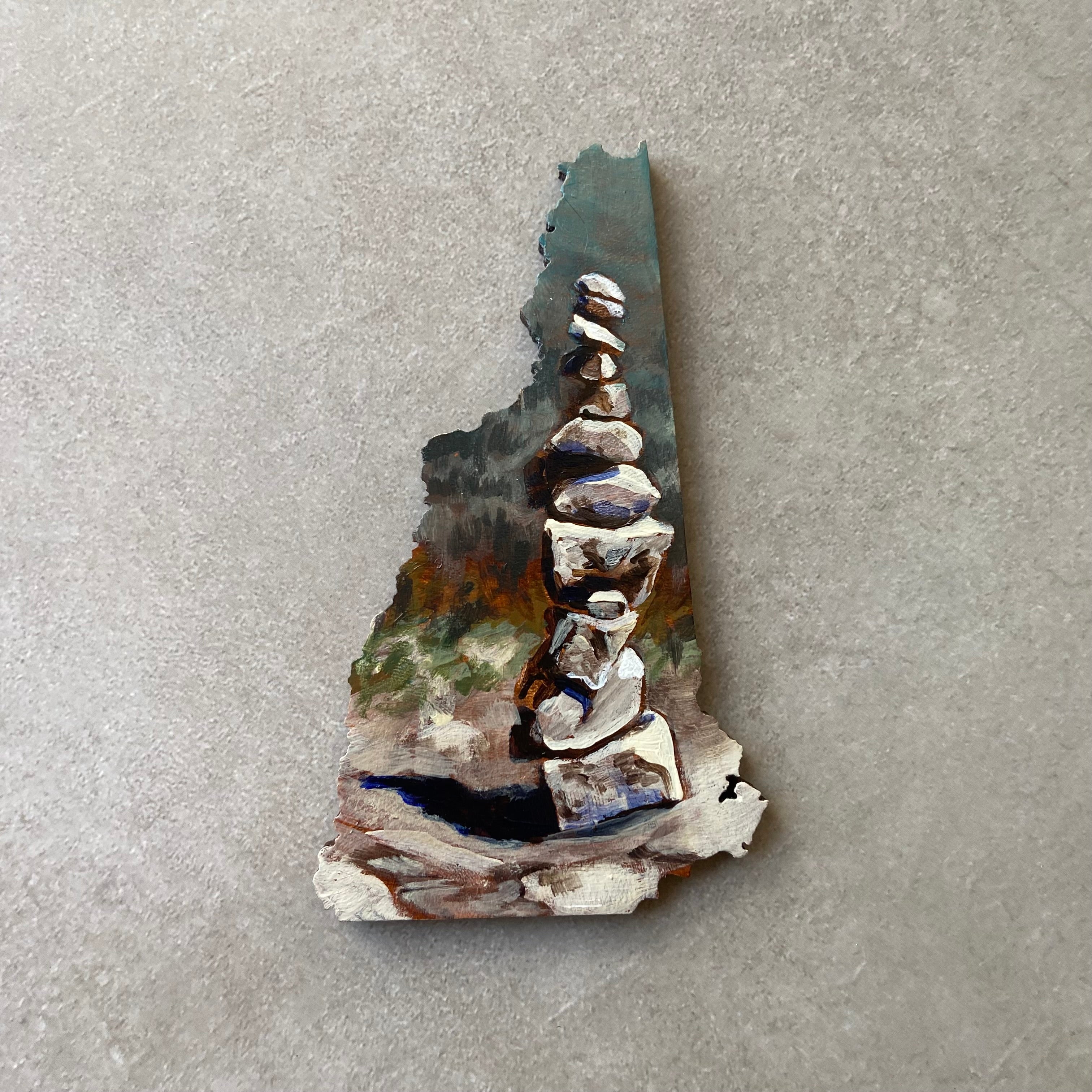 A New Hampshire-shaped painting of stacked rocks on a rock surface, with implied forest painted in the distance. 