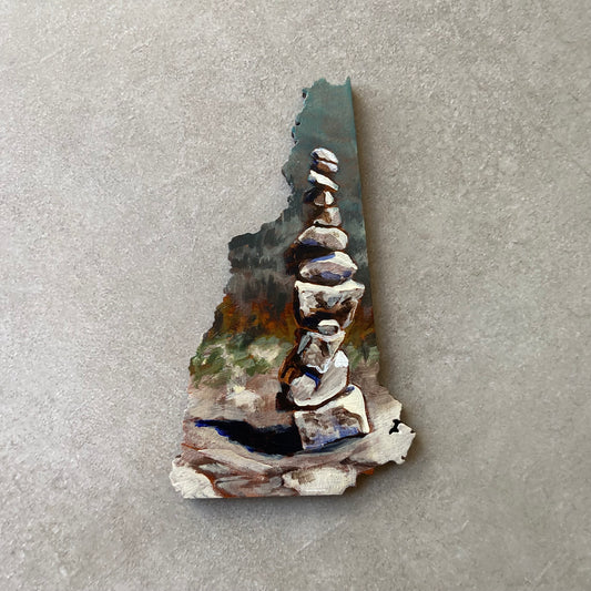 A New Hampshire-shaped painting of stacked rocks on a rock surface, with implied forest painted in the distance. 