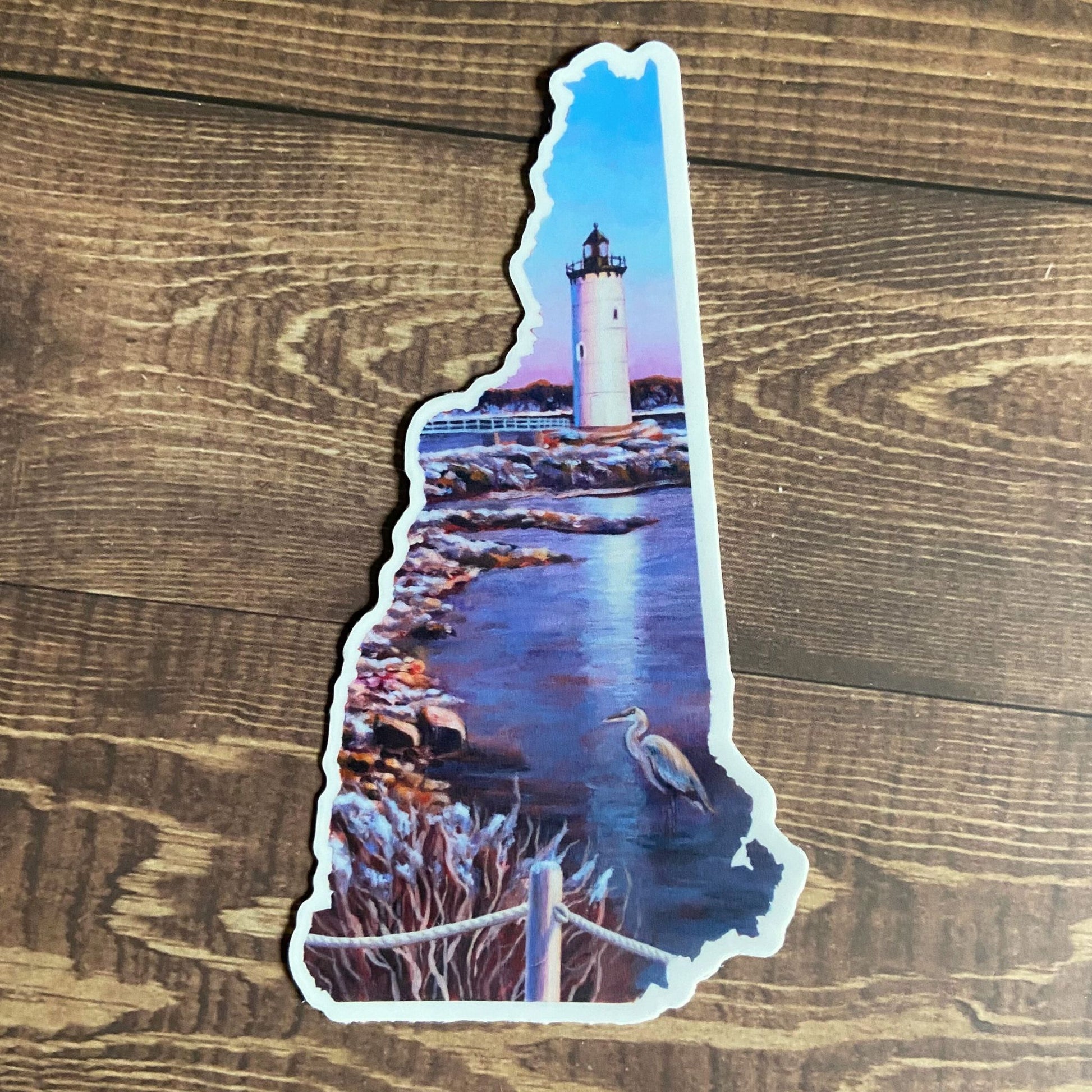 A vinyl sticker depicting a painted lighthouse scene with a heron, in the shape of New Hampshire. The sticker is mainly blue. 