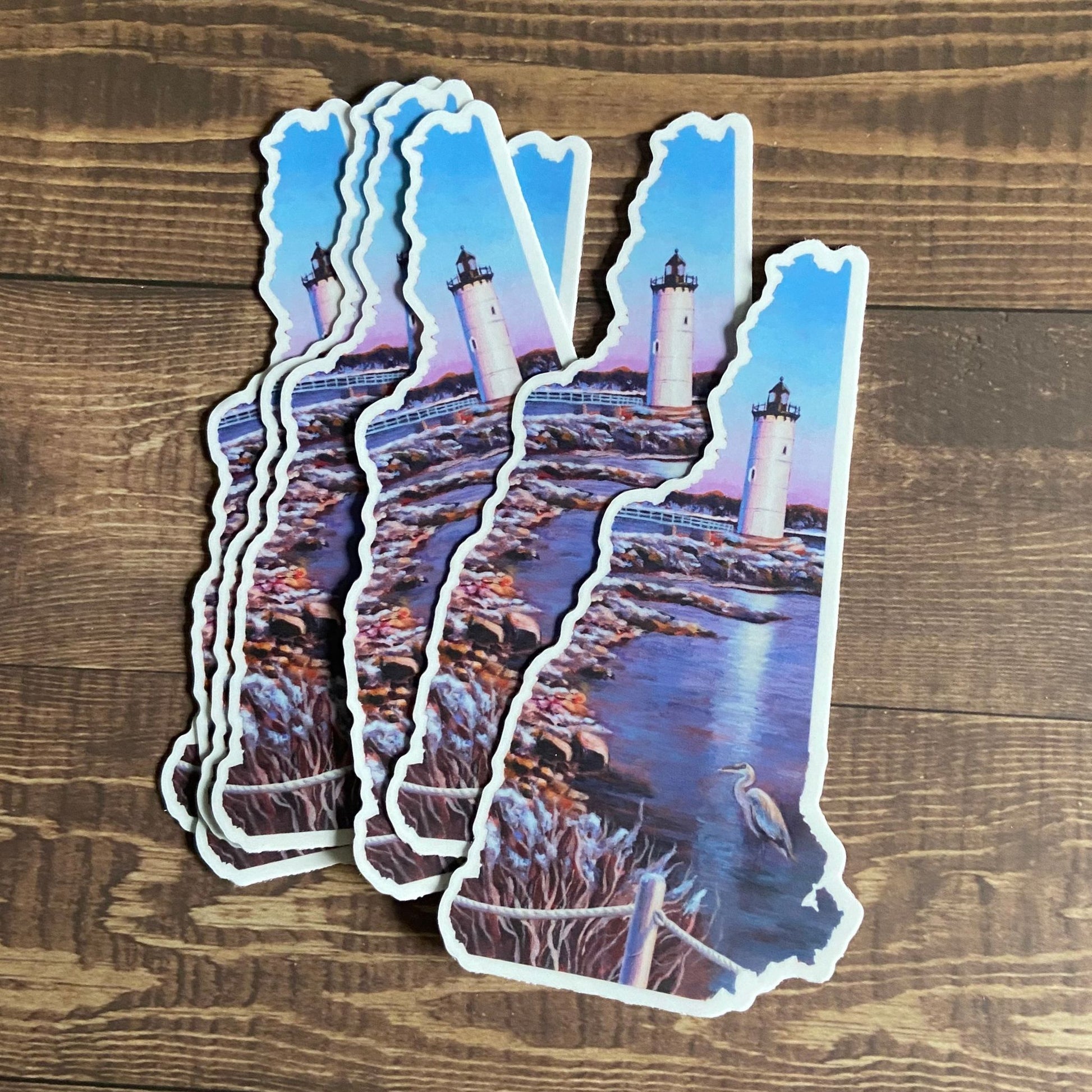 A small pile of stickers depicting a painted lighthouse scene with a heron, in the shape of New Hampshire. The stickers are mainly blue. 
