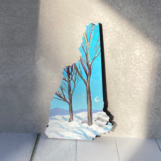 A New Hampshire-shaped painting of two bare-branched trees in the snow with mountains in the distance. The sky is clear, blue, and has a crescent moon at right. The painting is propped up in a patch of sunlight.