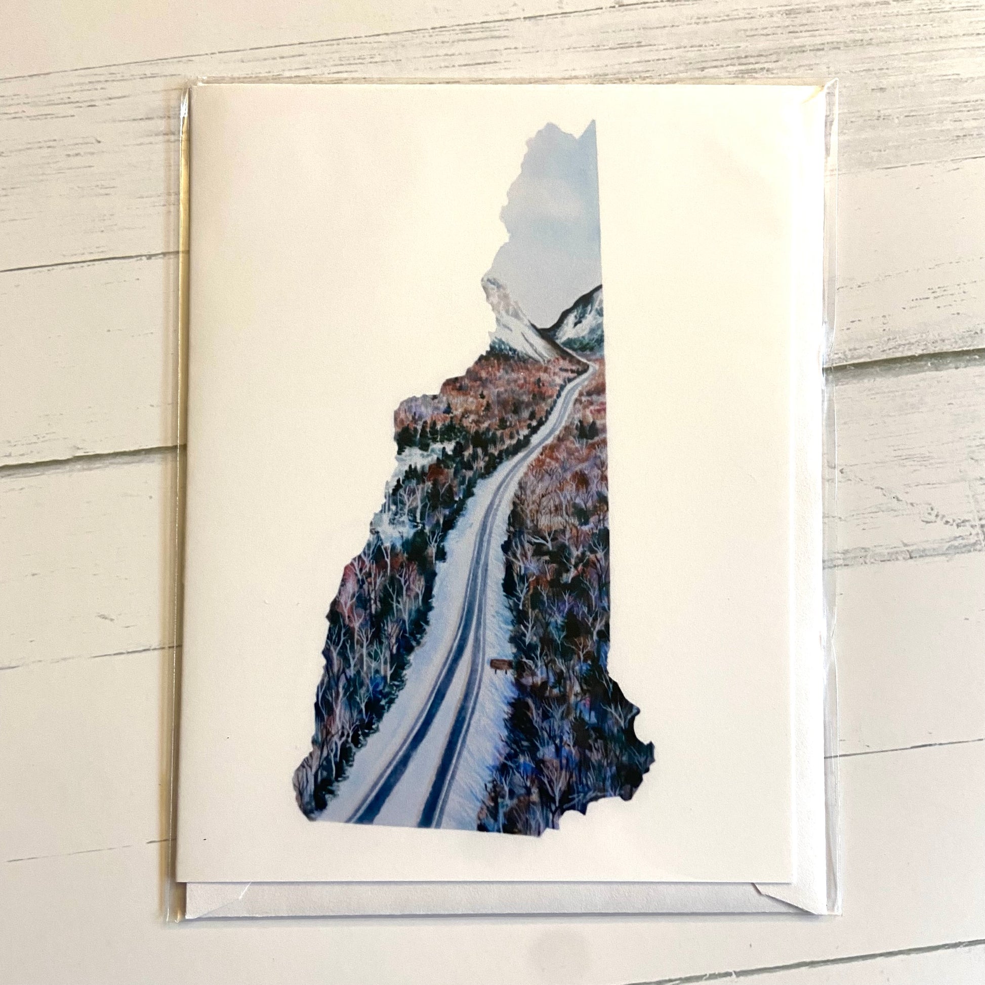 A folded white greeting card with an image in the shape of New Hampshire showing a snowy Route 93 in Franconia Notch. 