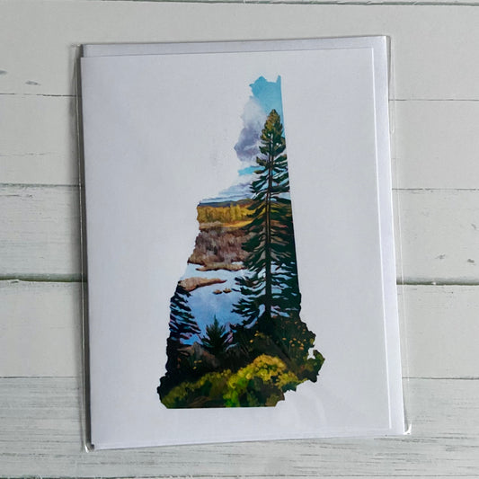 An overhead view of a greeting card with my painting, New Hampshire IV, printed on the front. Depicted is a painting in the shape of the state of New Hampshire with a tall pine tree in front of a marsh and fall foliage. The image is on a white background. 