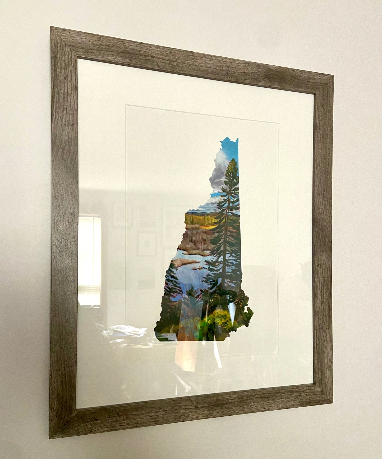 A painting of a fall landscape in the shape of New Hampshire, framed on a light gray wall. The painting depicts a tall pine tree in front of a marsh and fall foliage. Around it is white. The frame is a wood-textured cool medium brown with a white mat. 