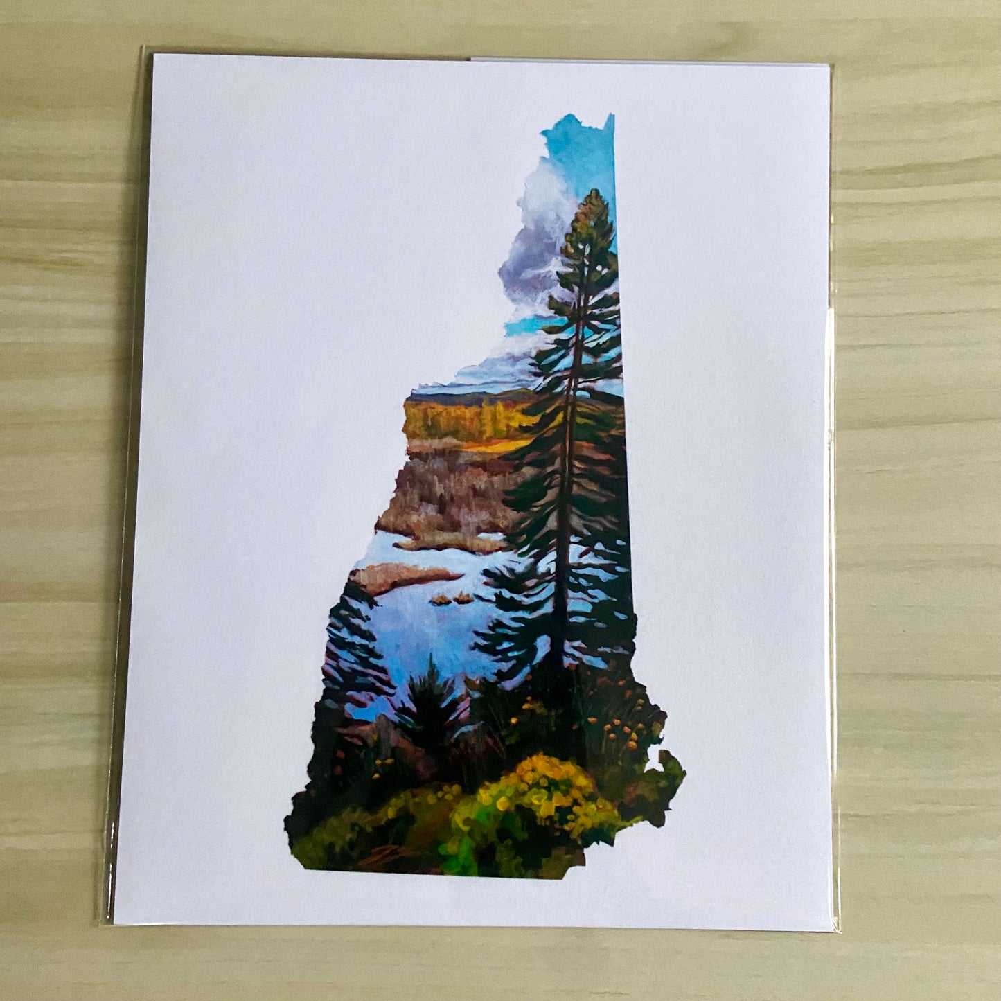 An overhead view of a print of my painting, New Hampshire IV. Depicted is a painting in the shape of the state of New Hampshire with a tall pine tree in front of a marsh and fall foliage. The image is on a white background. 