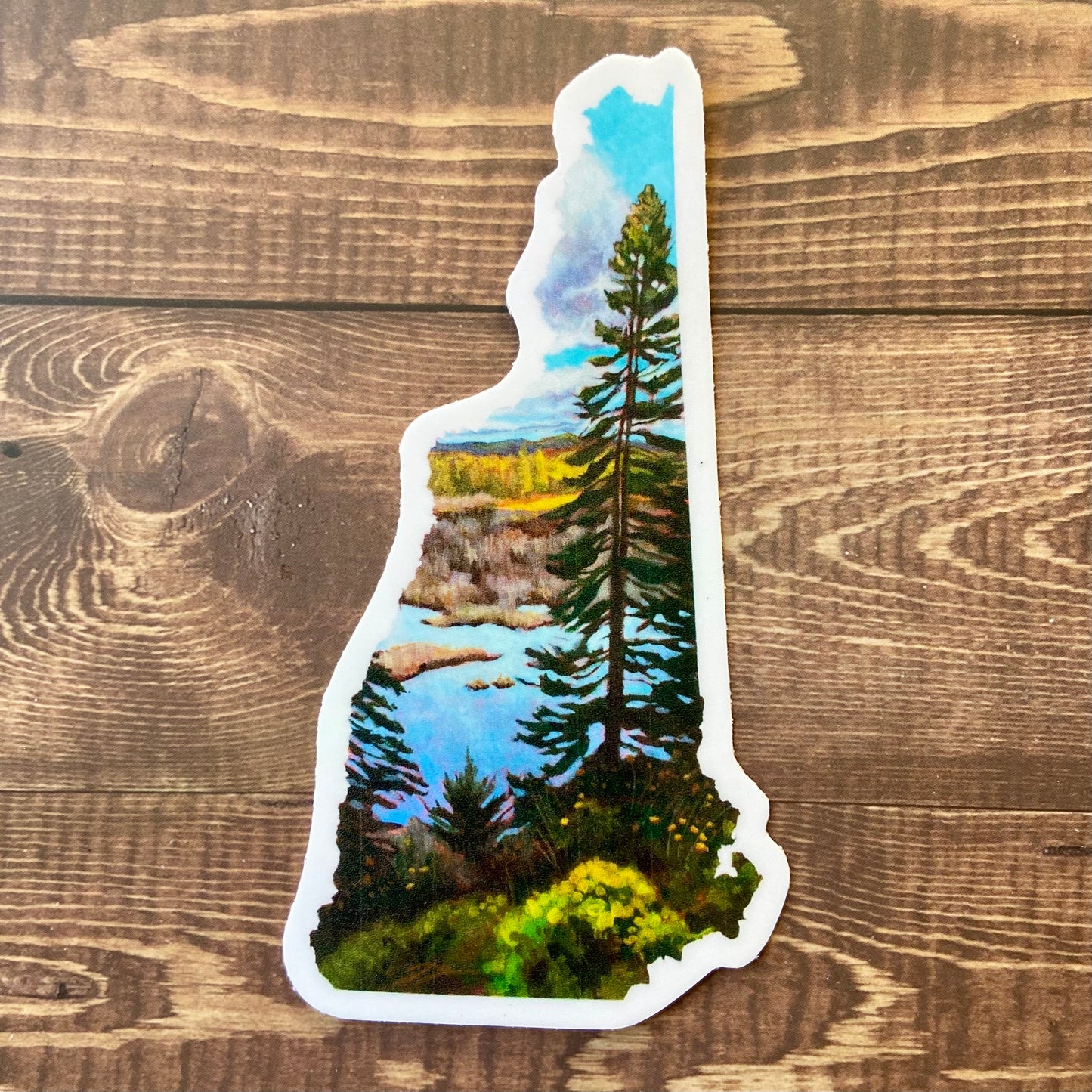 A sticker in the shape of New Hampshire depicting a tall pine tree in front of a marsh on a sunny fall day. 