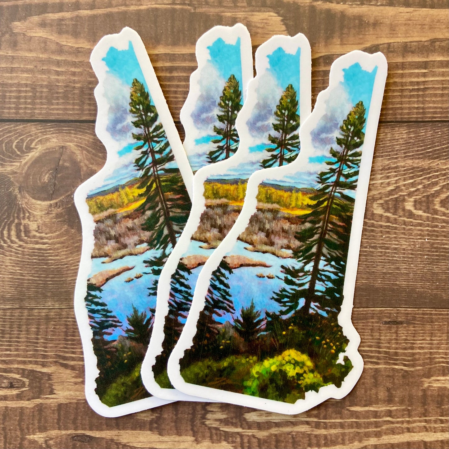 A small pile of stickers in the shape of New Hampshire depicting a tall pine tree in front of a marsh on a sunny fall day. 