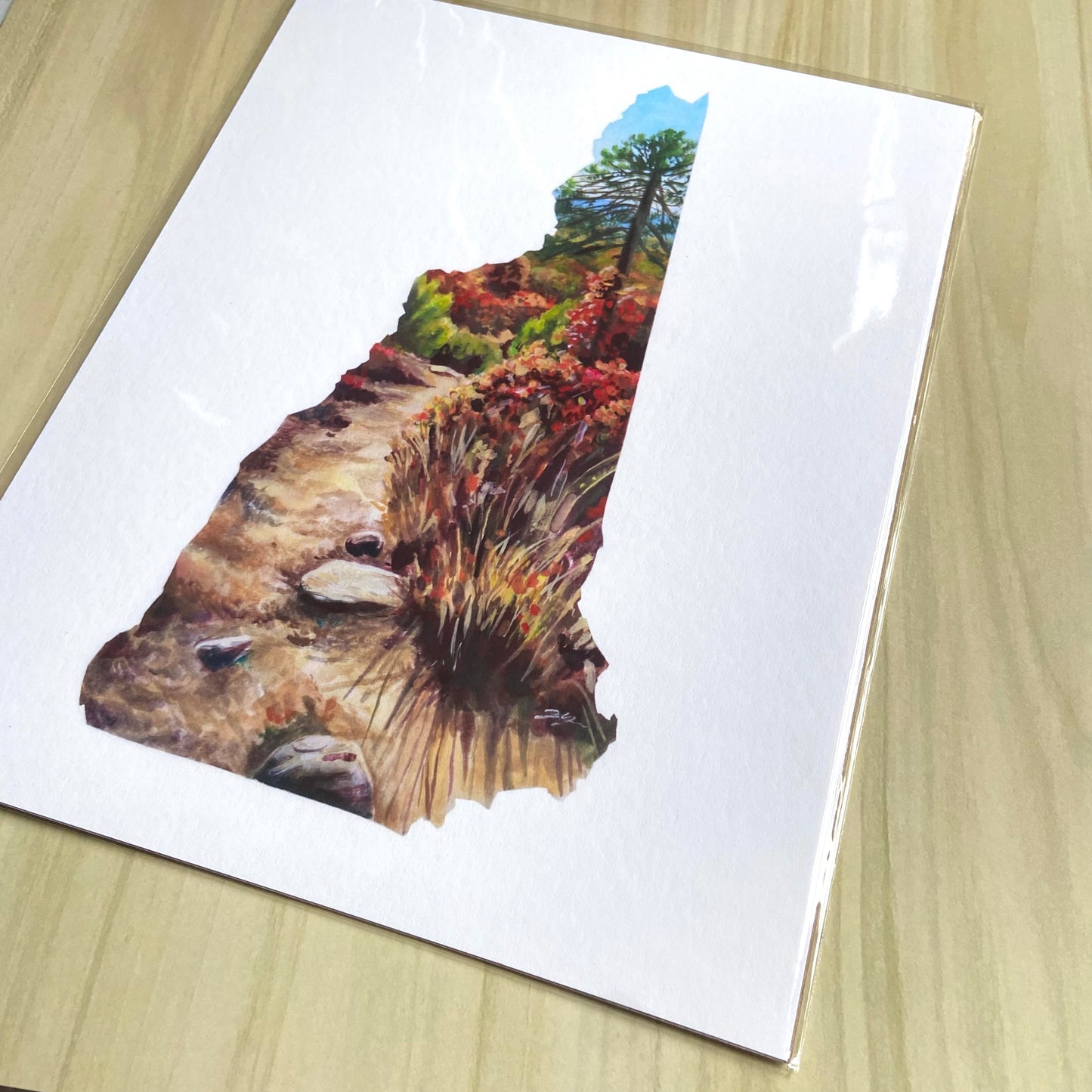 A detail view of the NH shaped art print. This is a copy of a painting showing a trail in the woods on a sunny day, within the map outline of New Hampshire. Nature is showing autumn colors.