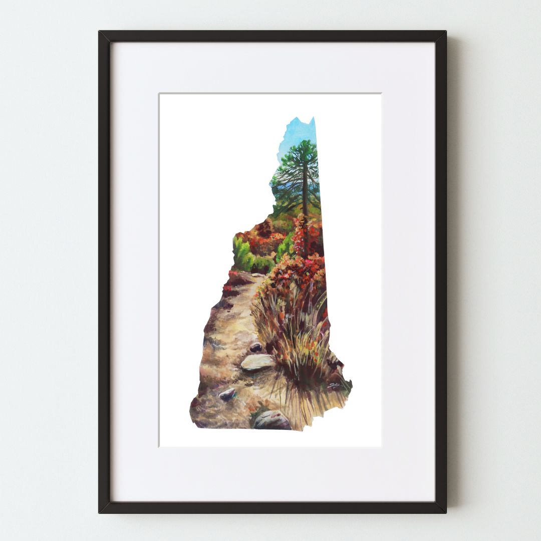 A mockup of the New Hampshire shaped landscape art print in a black frame with a white mat. The painting shows a dirt path through brush to a pine tree and fall foliage under a blue sky.