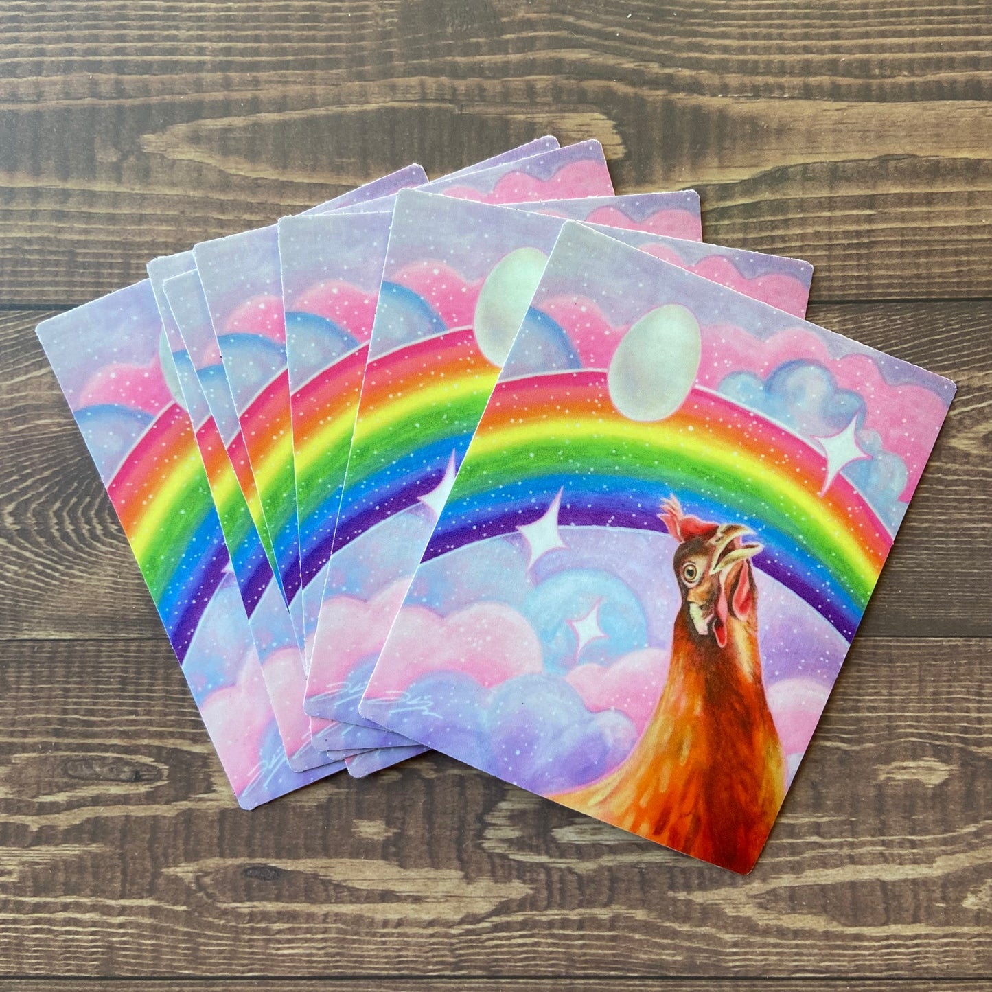 A small pile of rectangular stickers of my painting "Nostalgia Hen", depicting an orange hen looking awestruck at a rainbow in a pink and purple sky. The are sparkles everywhere and an egg floating overhead. 