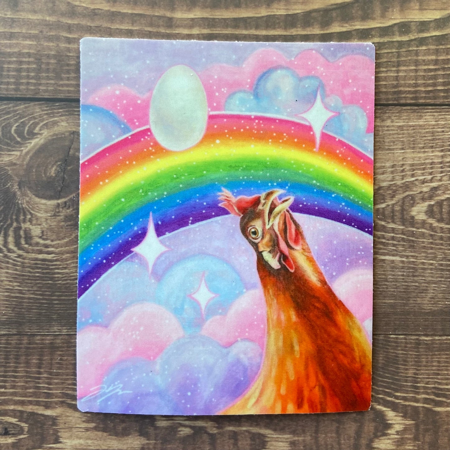 A rectangular sticker of my painting "Nostalgia Hen", depicting an orange hen looking awestruck at a rainbow in a pink and purple sky. There are sparkles everywhere and an egg floating overhead. 