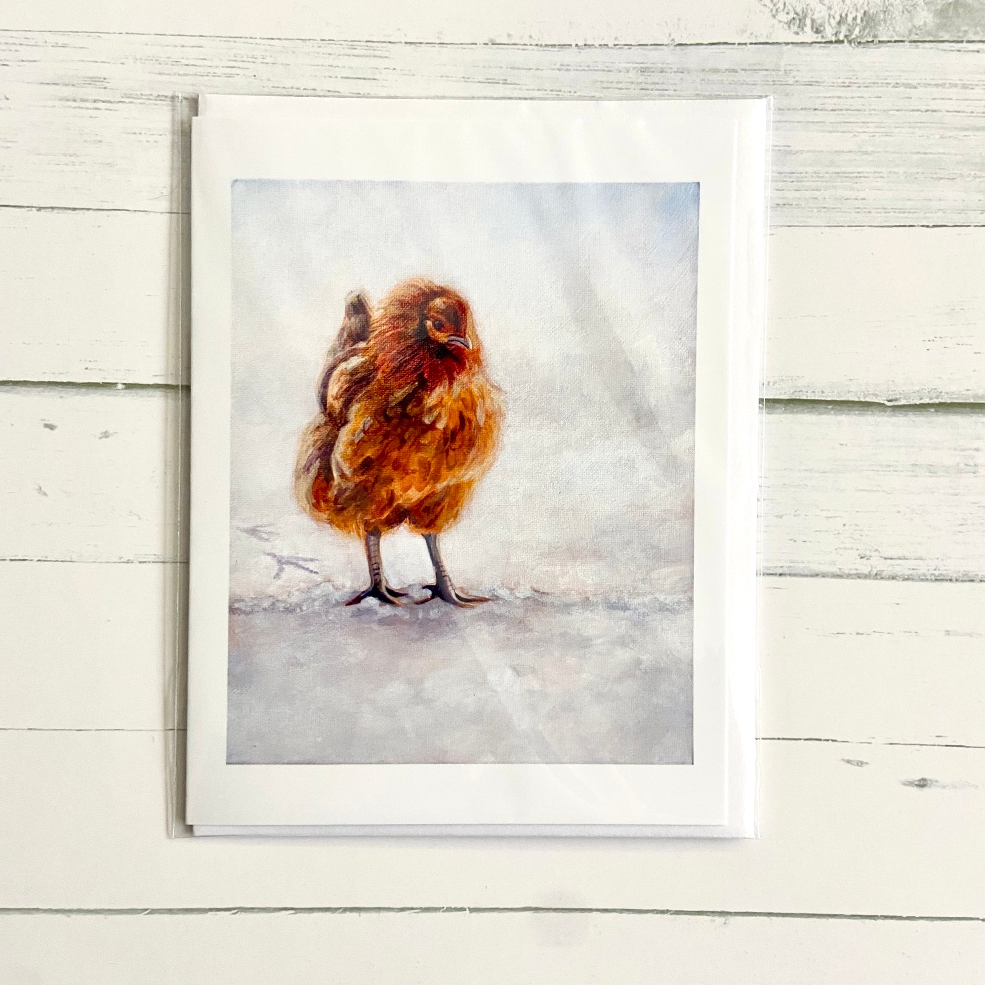 A folded white greeting card with a painting on the front of an orange chicken standing in snow. 