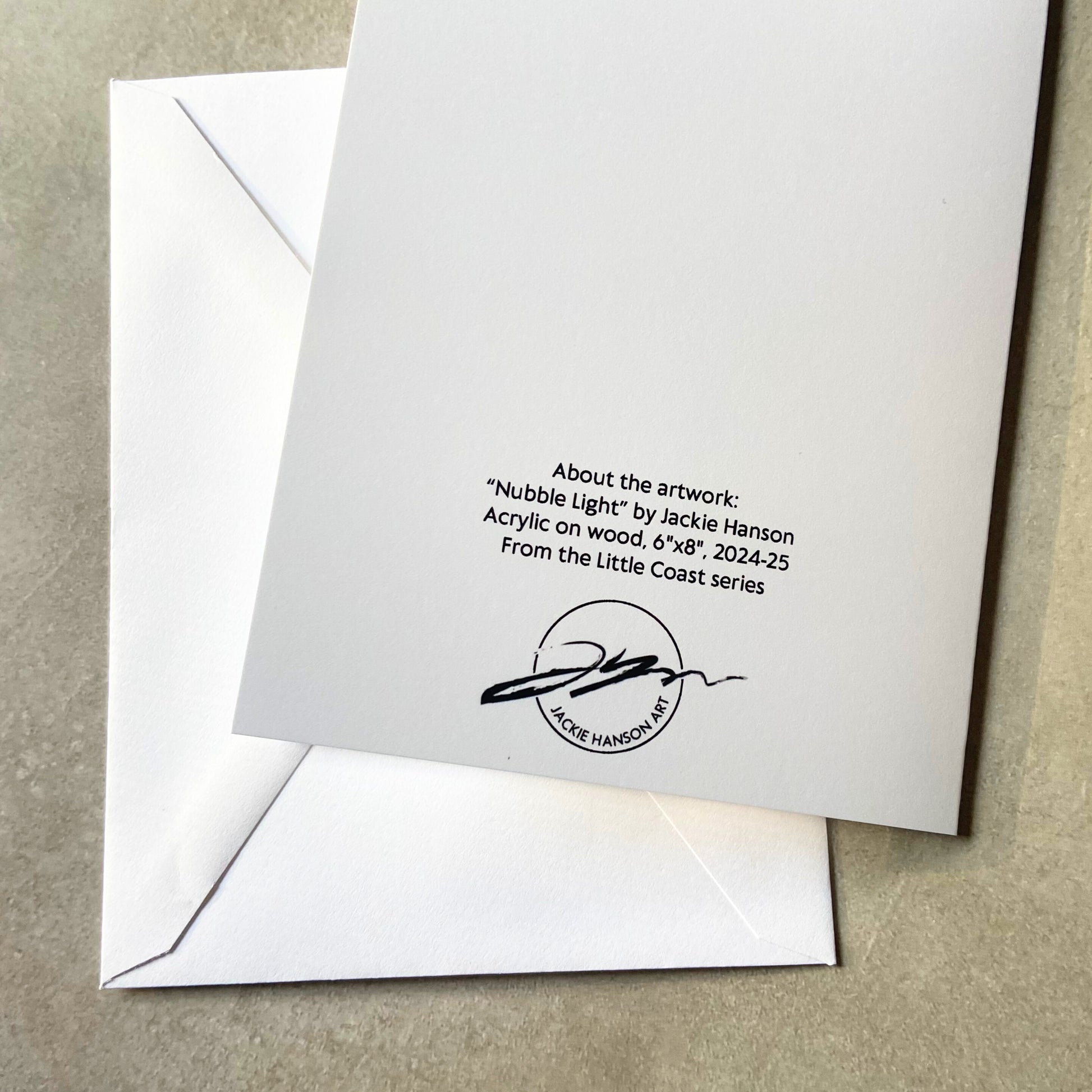 A folded white greeting card, face-down, and envelope on a grey surface. On the back of the card reads, "About the artwork: "Nubble Light" by Jackie Hanson. Acrylic on woodm 6"x8", 2024-25. From the Little Coast series." Below is the artist's signature/logo.