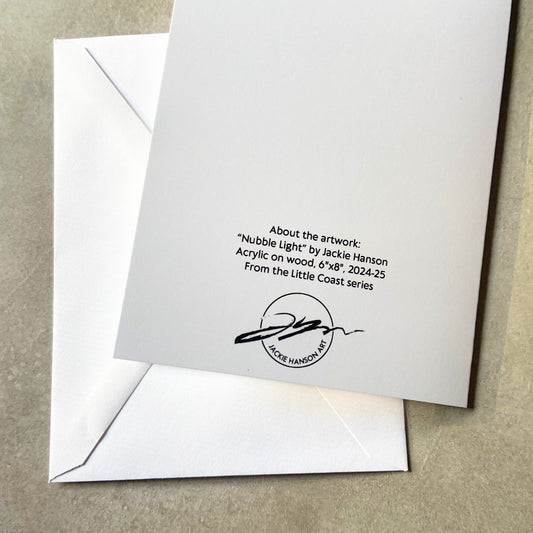 A folded white greeting card, face-down, and envelope on a grey surface. On the back of the card reads, "About the artwork: "Nubble Light" by Jackie Hanson. Acrylic on woodm 6"x8", 2024-25. From the Little Coast series." Below is the artist's signature/logo.