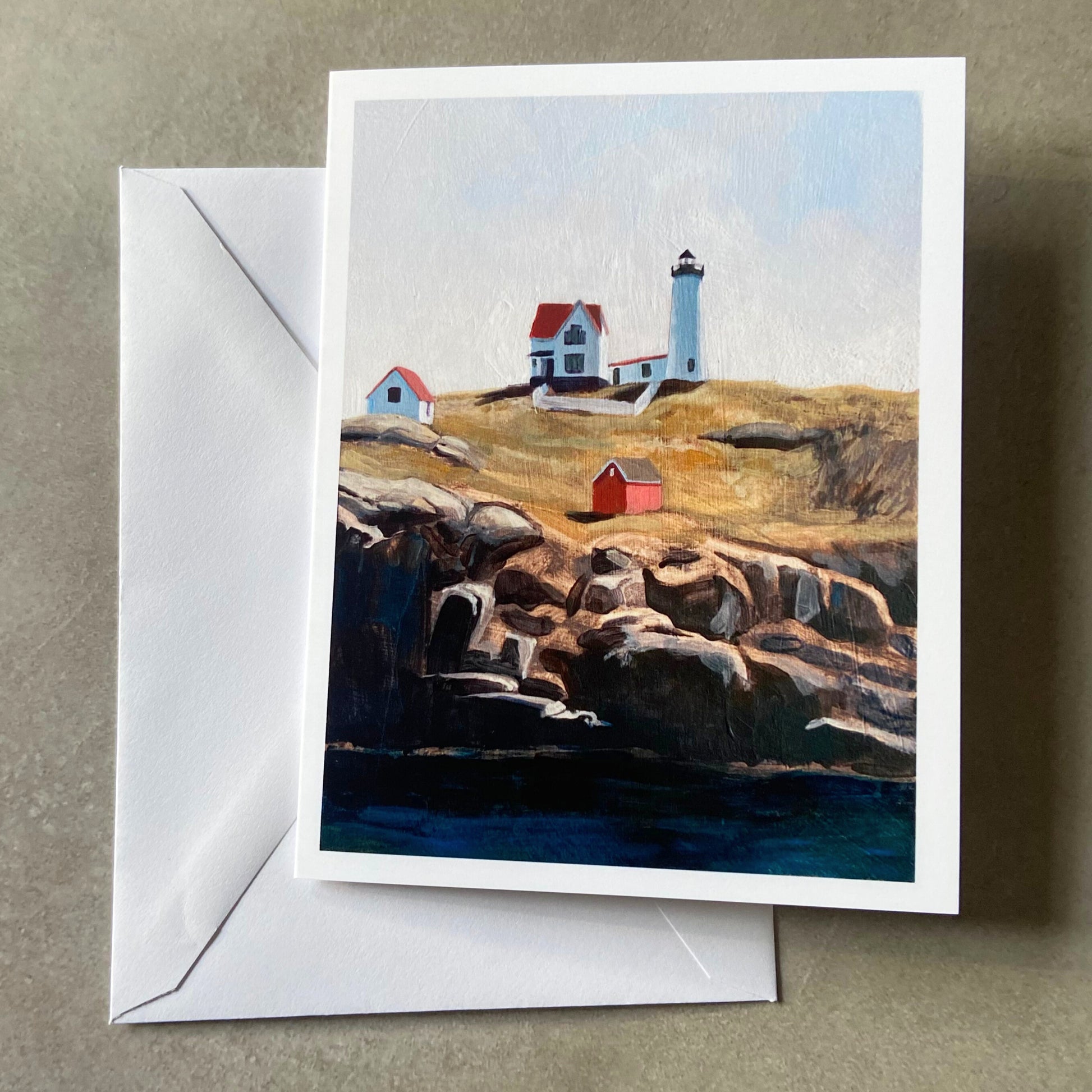 A folded white greeting card and envelope on a grey surface. Depicted on the card is a painting of Nubble Lighthouse in York, Maine, standing on a tall island with rocky cliffs in front above deep blue water. 
