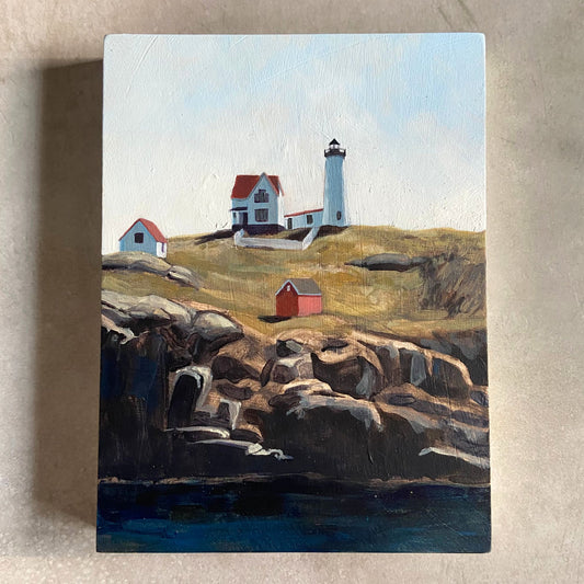 An acrylic painting on wood panel sitting on a beige, textured background. The artwork, portrait-oriented, shows a lighthouse on a green piece of land under a blue sky. The land ends in rocky cliffs with deep blue water below. 