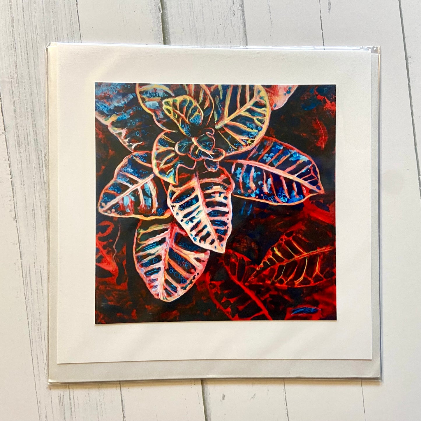 A square, folded white greeting card with an image of my painting, Out of Darkness. Shown is the houseplant Croton in brilliant primary colors. The background is red and black leaves. 