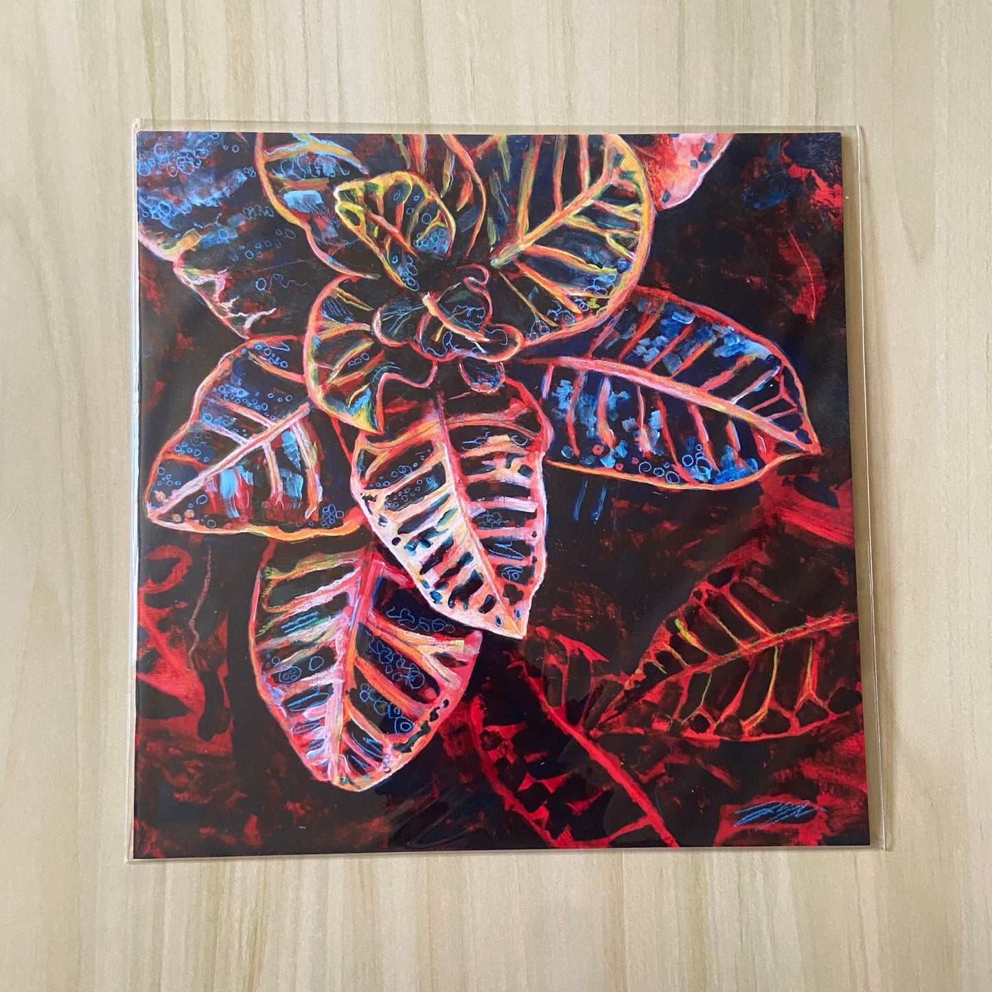 A square art print of a variegated Croton houseplant. With a vibrant red base and dark shadows, the painting seems to glow. The patterned leaves show bright, playful pastel colors amidst all the black. Great moody yet colorful decor for plant lovers.