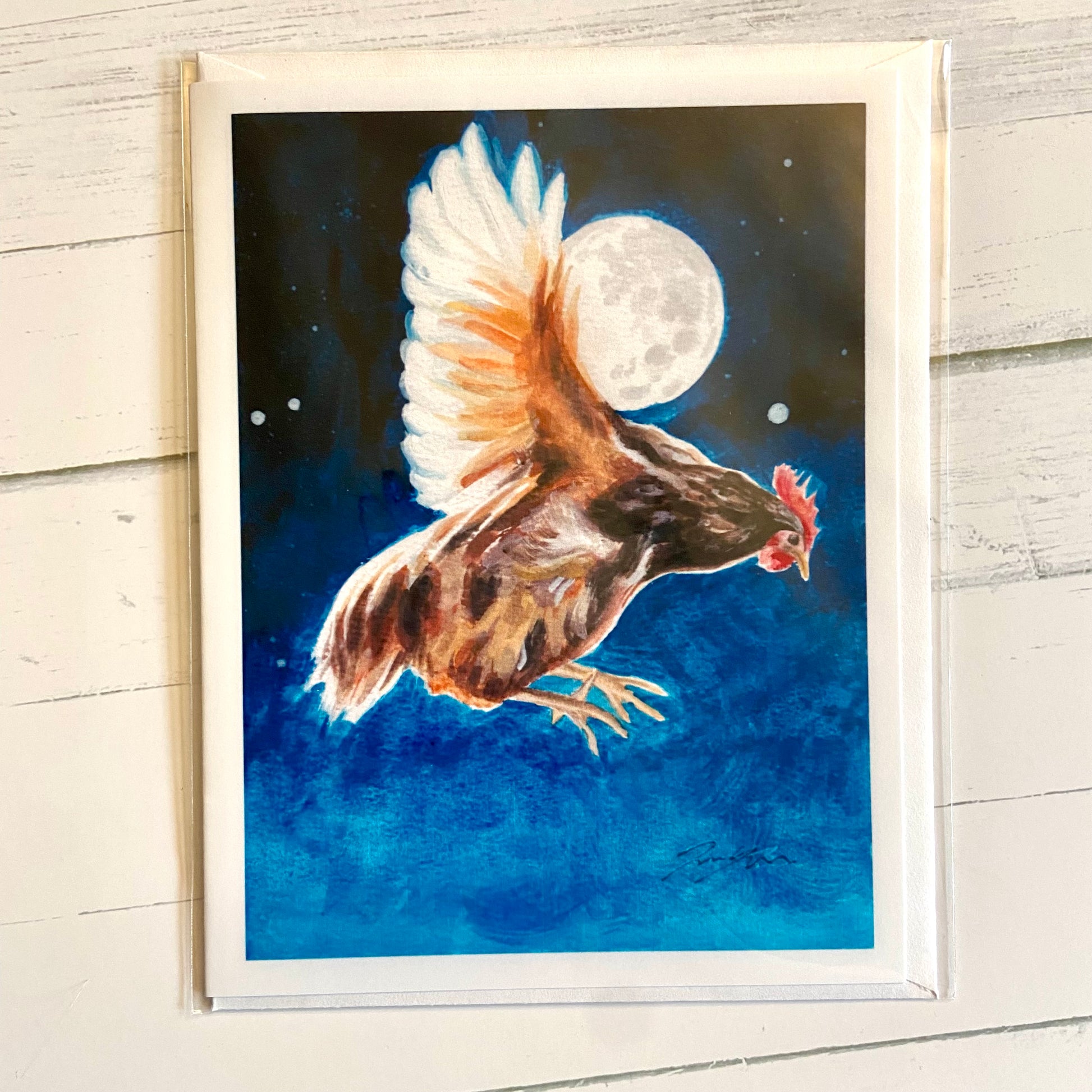 A folded white greeting card with a painting on the front of a chicken in flight before a night sky, its wing partially eclipsing a full moon. 