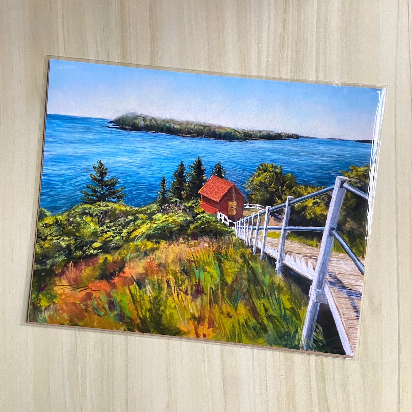 An art print of a coastal Maine landscape painting made with soft pastels. Multicolor abstract grasses are in the foreground, with a path downhill to a red shed. In the background is the ocean and sky with a forested island in the distance. 