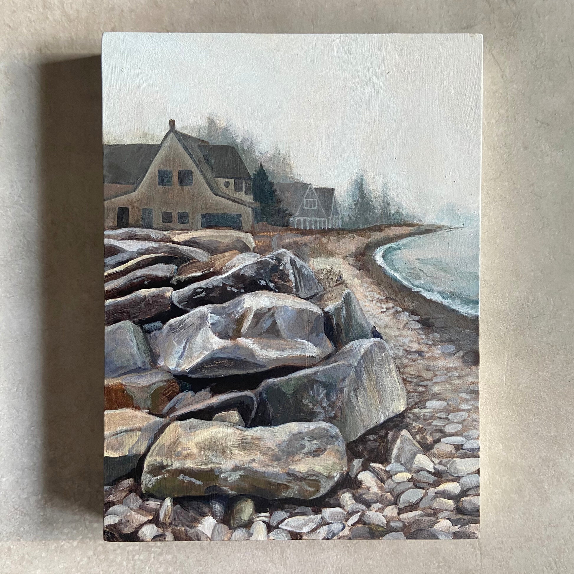 An acrylic painting on a wood panel, sitting on a beige, textured background. Depicted in the portrait-oriented artwork is a pile of sea-weathered rocks on a graveled beach. There is a row of beachfront homes behind the rocks, fading into the thick fog with distance. 