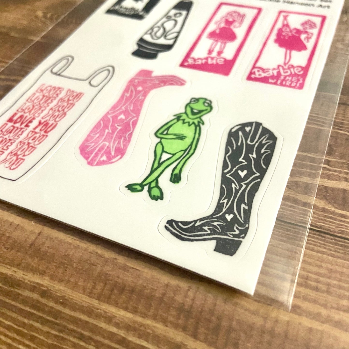 A close-up, angled view of my eraser art sticker sheet "POP! cultured". Individual cut lines around the 8 stickers can be seen. The stickers are around 1x2 inches each and include Barbies, Kermit the Frog, cowboy boots, a lava lamp, and more. 