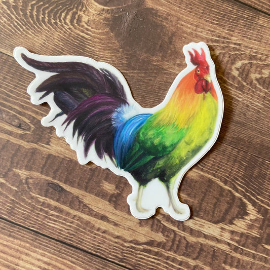 A sticker of a rainbow-feathered rooster in profile sitting on a wooden surface. The decal is die-cut to shape with a thin white border. The chicken has a red comb with the rest of the rainbow extending across his body to dark purple tail feathers.