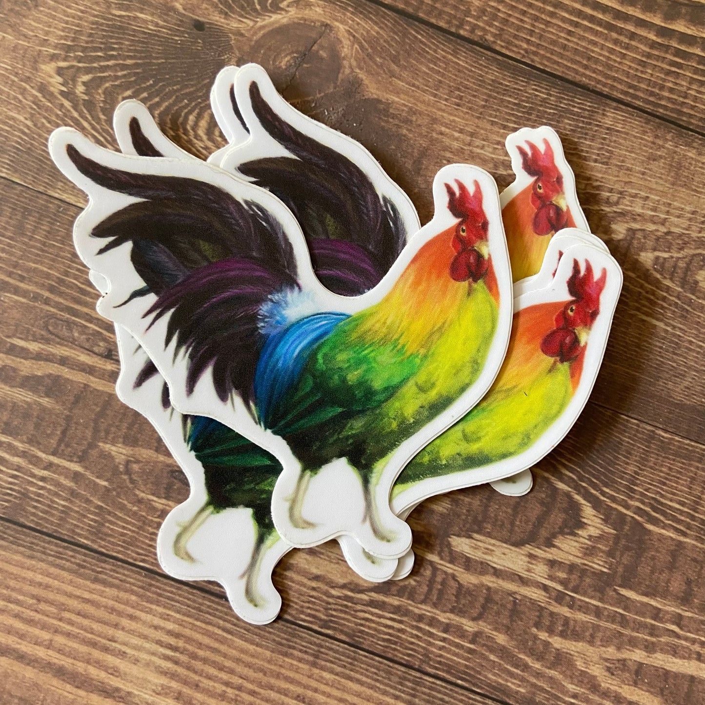 A small pile of rainbow rooster stickers on a wood surface. The birds are standing facing right, showing off their vibrant multicolor plumage. Each decal is die-cut to shape with a narrow white border.