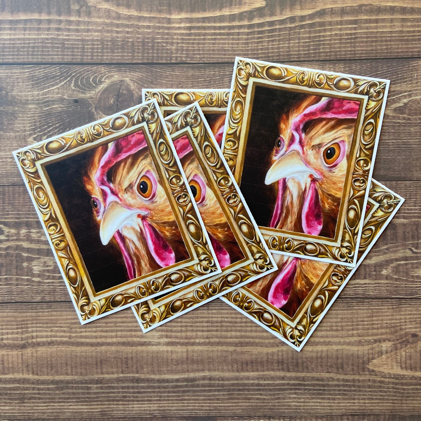 A small pile of stickers of my painting, "Regal Chicken" or "Hennifer", depicting a hen's angry face inside an ornate gold frame (also painted.) 