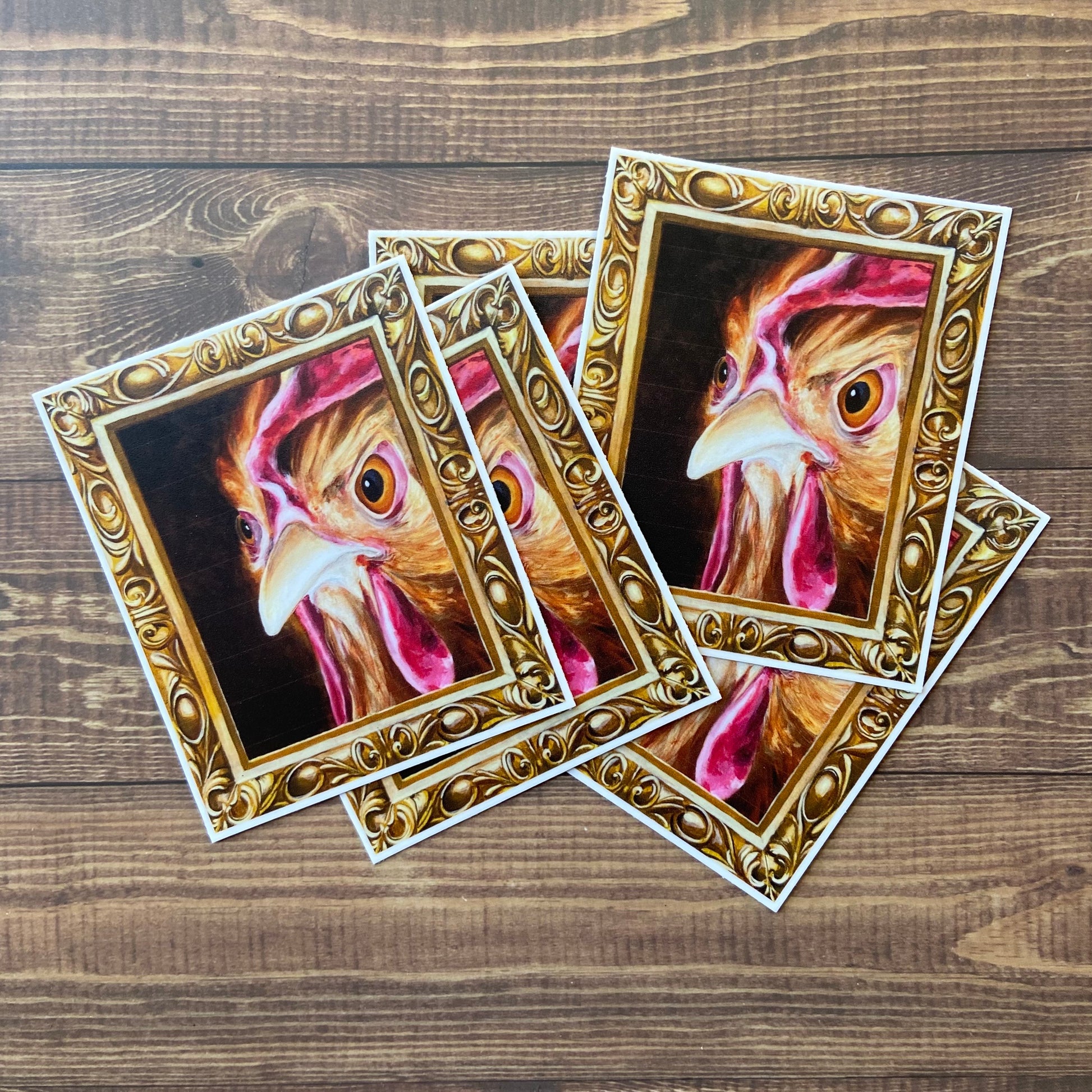 A small pile of stickers of my painting, "Regal Chicken" or "Hennifer", depicting a hen's angry face inside an ornate gold frame (also painted.) 