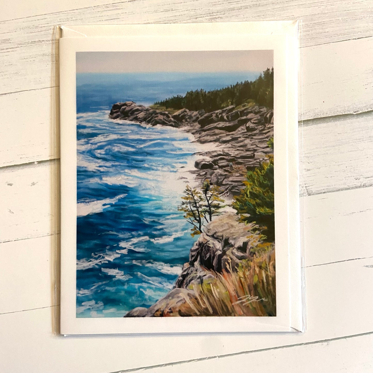 A folded white greeting card with a painting of a rocky Maine cliff and beach meeting the bright blue sea. 