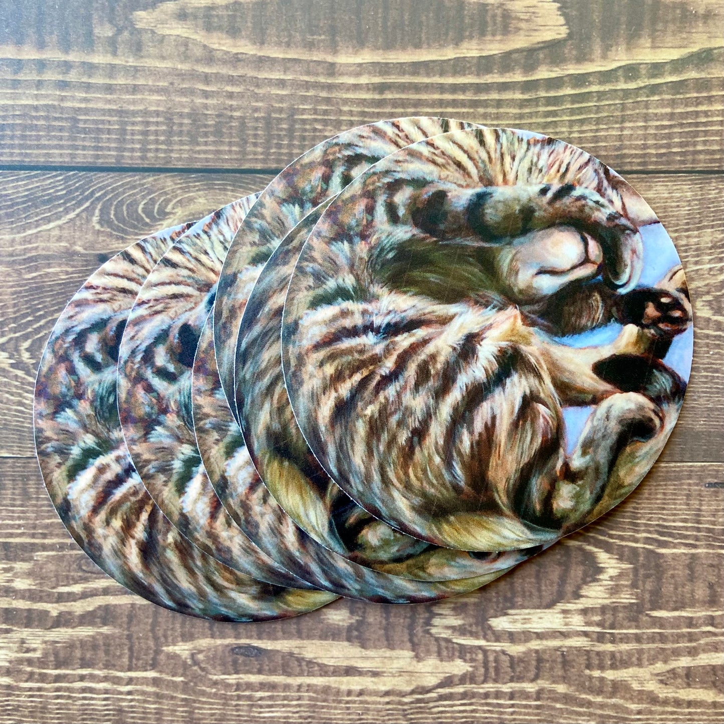 A small pile of stickers of my painting "Roundcat" depicting a gray/brown striped tabby cat curled in a circle sleeping. Her paw is covering her eyes so only her nose and mouth are showing, with a slight content smile on her face.