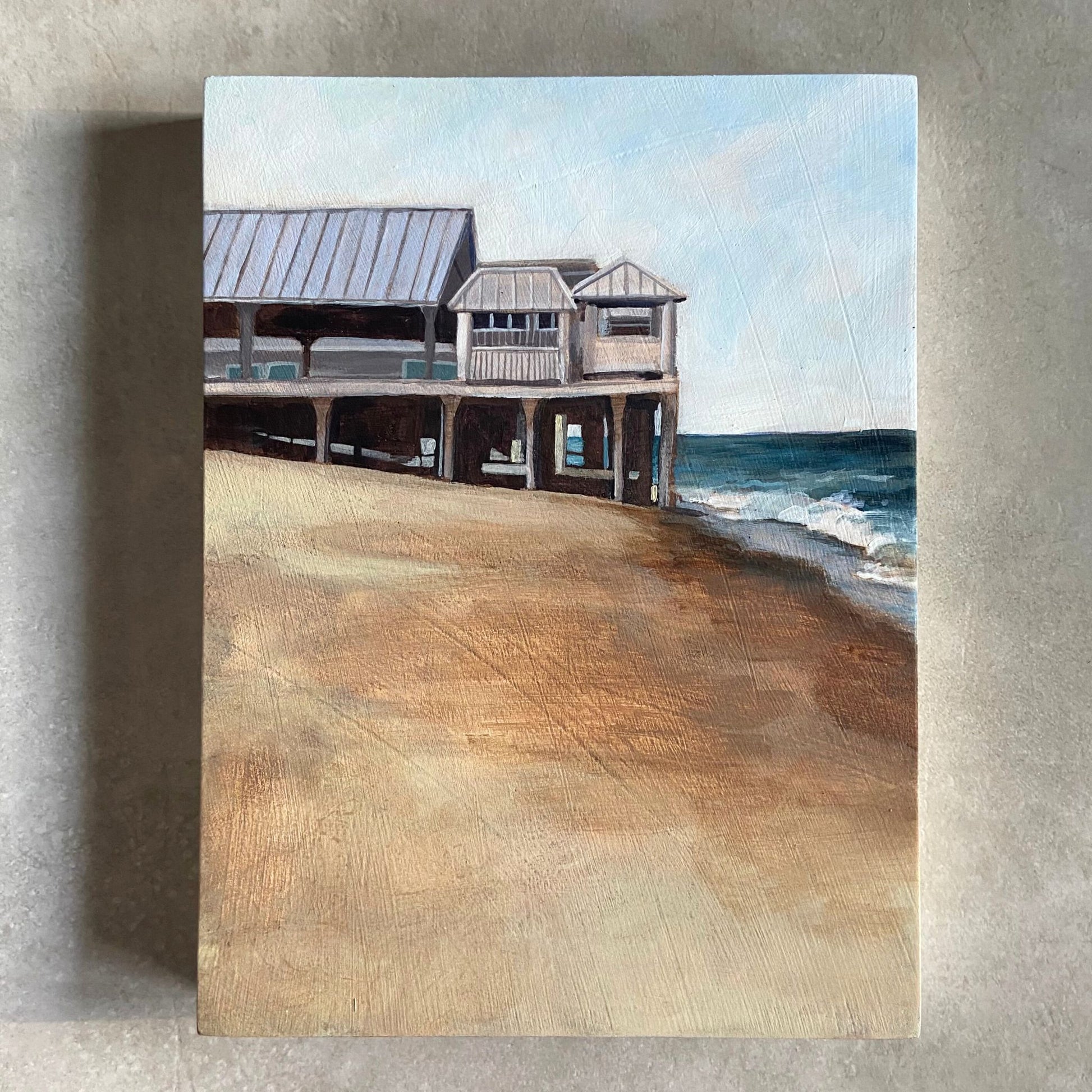 A beach painting on wood panel, sitting on a beige, textured background. The artwork, portrait-oriented, shows a boardwalk with light colored buildings in the top half of the painting, extending out onto the beach to meet the shore at right. The bottom half of the painting is largely consumed by tan-colored sand, made with thin layers of paint that allow the panel's texture to show through. 