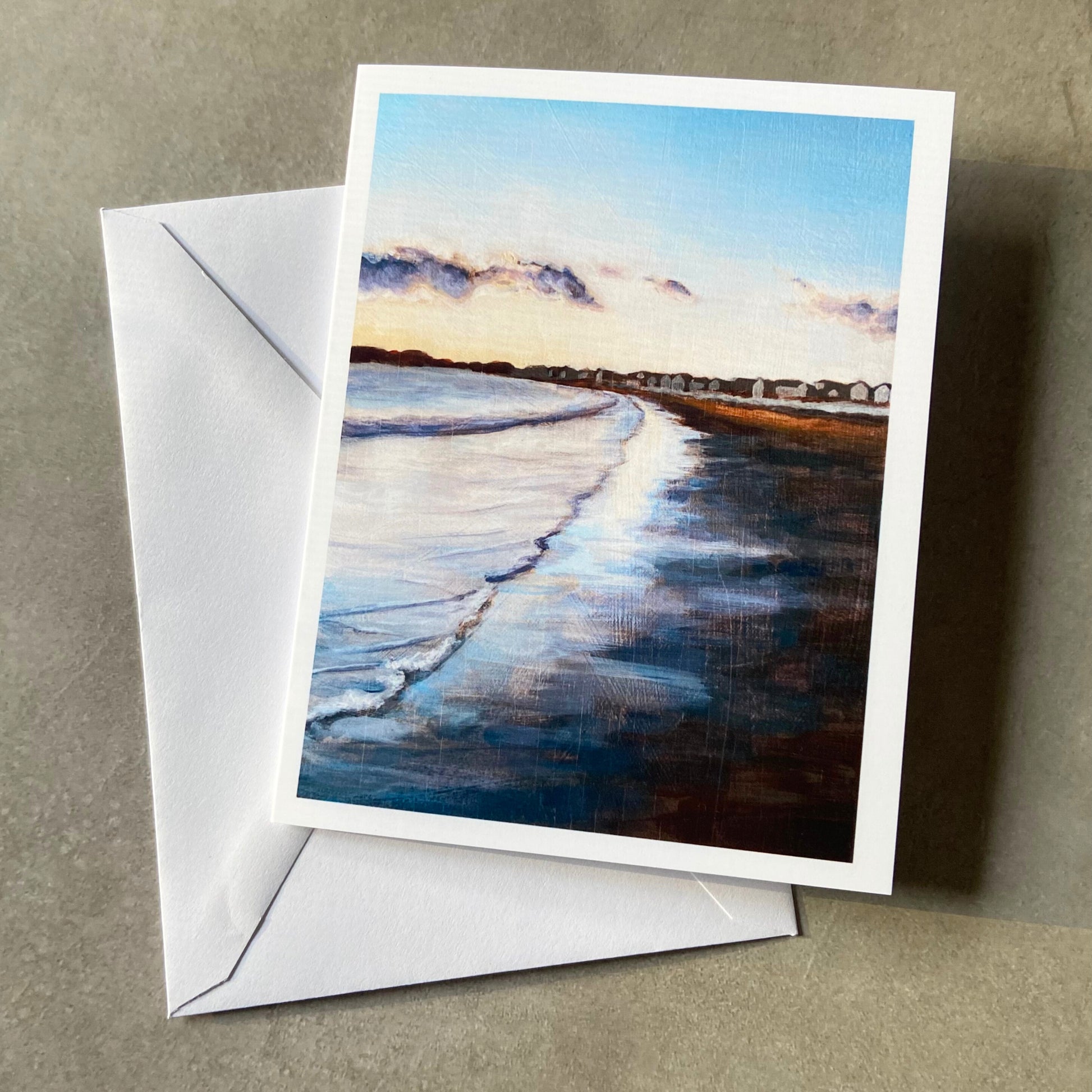 A folded white greeting card and envelope on a grey surface. The card shows a painting of shallow waves meeting sand at twilight hour, the sky reflecting strongly on the water. In the distance are a row of beach houses. 