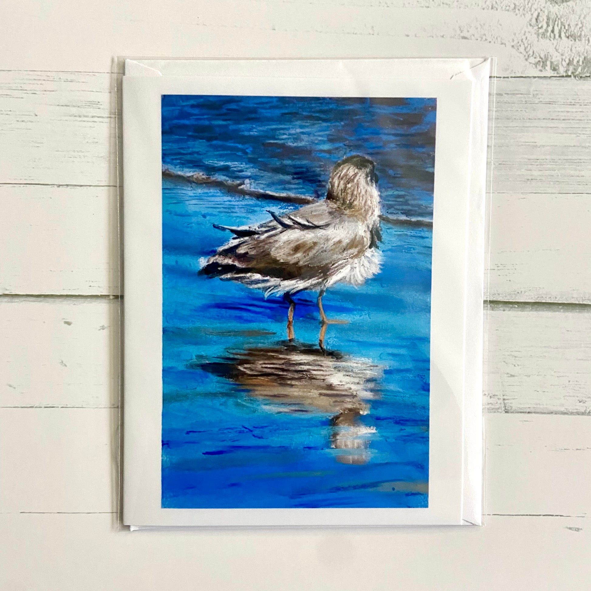 A folded white greeting card with a painting on the front of a seagull standing on wet sand, facing toward the waves. The reflection of blue sky and the bird make this painting interesting.