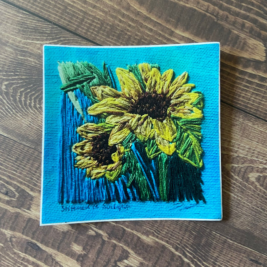 A square sticker of an embroidered painting of two sunflowers. The texture of the canvas and the texture of the thread are very clear. The painting is primarily light blue with the sunflowers in vibrant yellow.