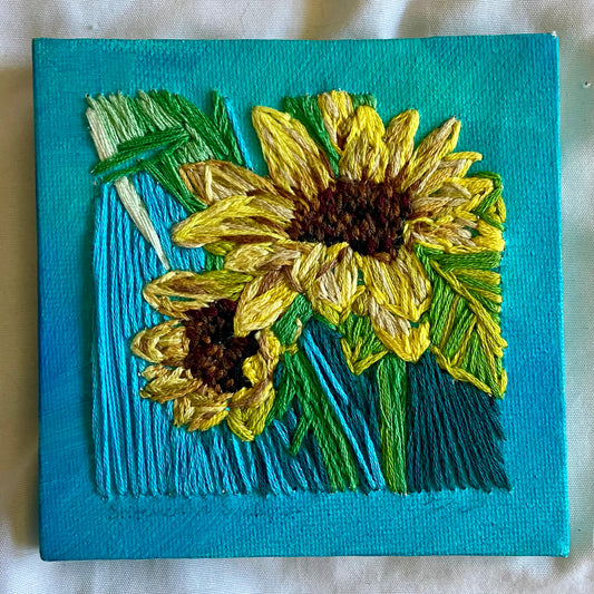 A small square painting made by embroidering two sunflowers onto a teal canvas. 