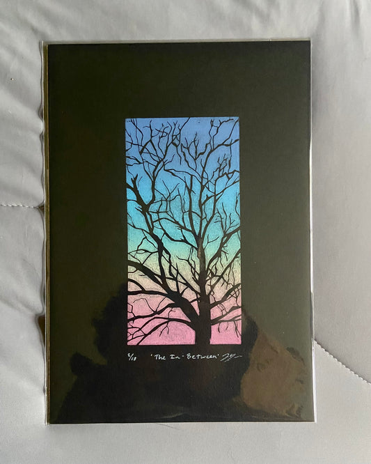 A vertically-oriented linocut print called "The In-Between" showing the branches of a tree carved away to reveal the black paper underneath. The negative space, or the sky, between branches is printed in a rainbow gradient. 