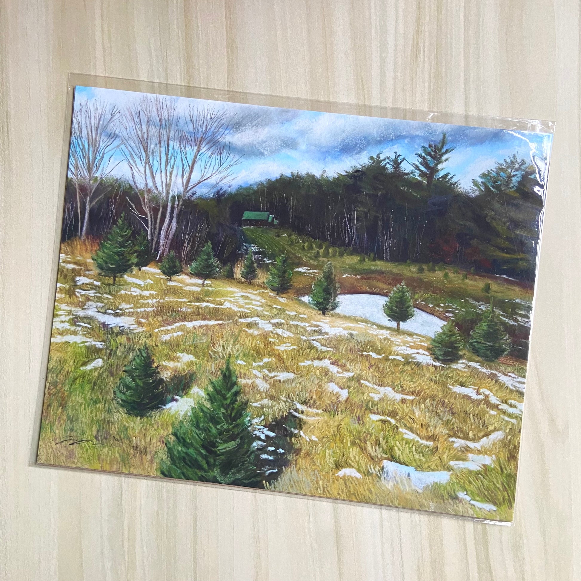 An art print of a pastel landscape painting. The artwork shows a dry hill dotted with snow and growing Christmas pines. Farther away is a frozen pond, more tree growth and a forest, a brown cabin with a green roof, all under a partly cloudy sky.