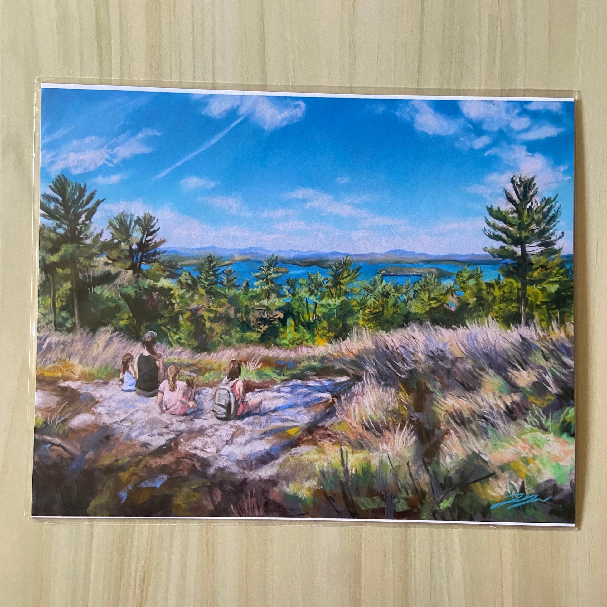 A paper print of a pastel landscape painting depicting a view from a mountain looking down at Lake Winnipesaukee. A mother and 3 young girls sit looking out at the view. The painting is primarily blue, with swaths of dry grass across the bottom.