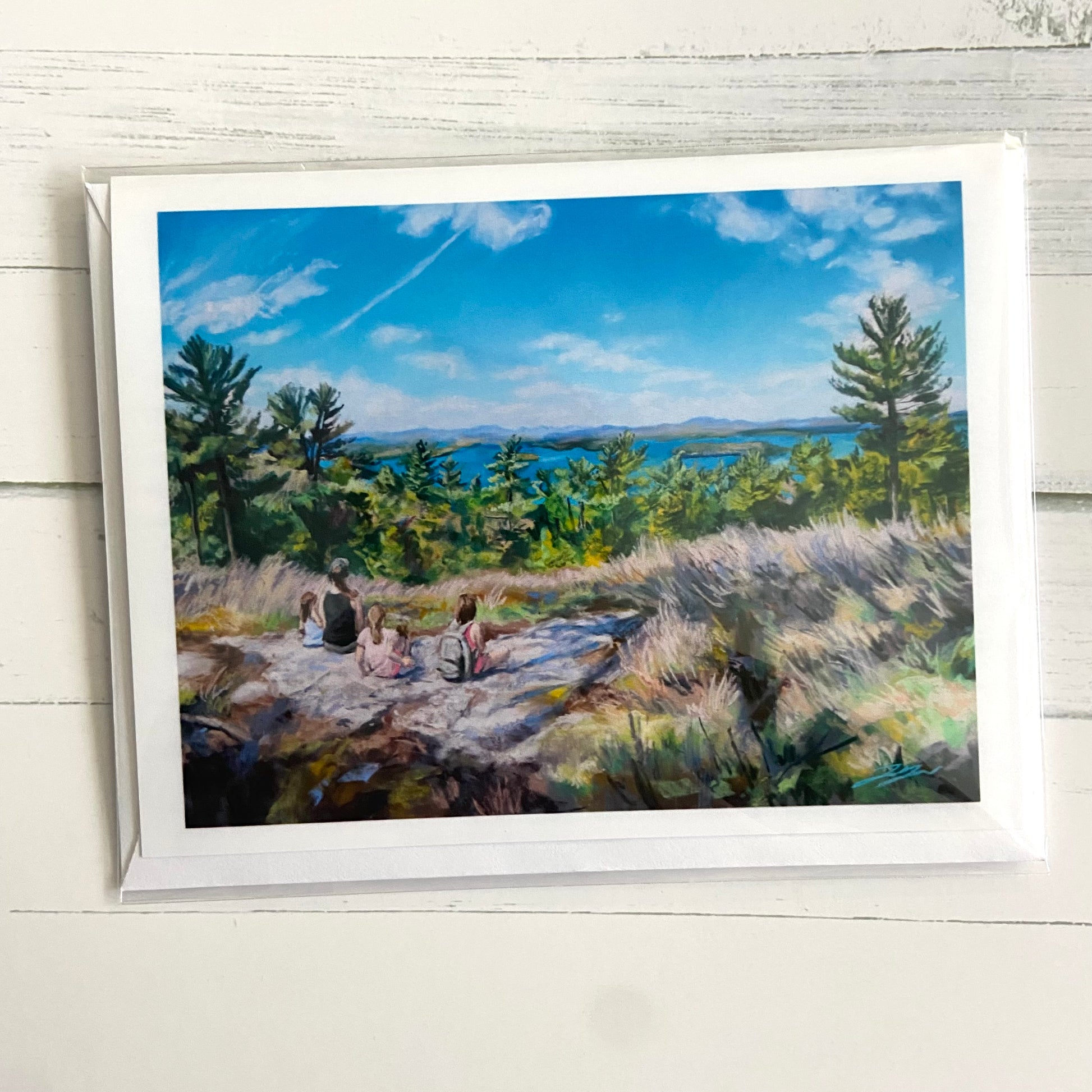 A folded white greeting card with a painting of the view at the top of a mountain, with Lake Winnipesaukee in the distance. There is a mom and three daughters sitting on the gorund looking at the view. 