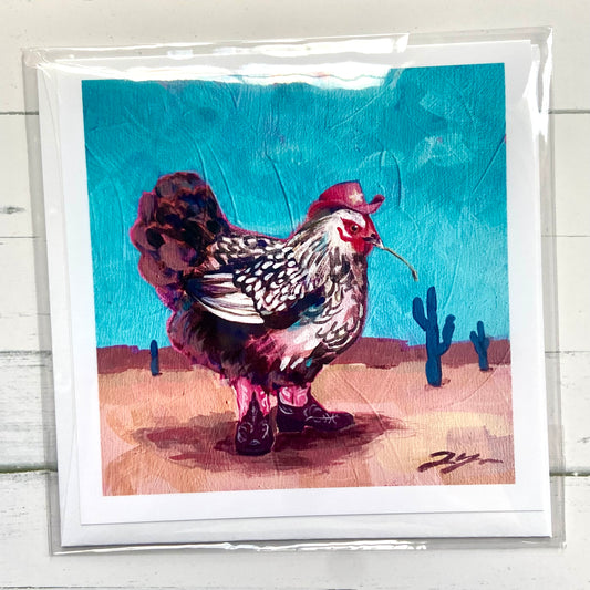 "Yeehaw" square greeting card by New Hampshire artist Jackie Hanson. A black and white feathered chicken wears a pink cowgirl hat and boots, standing in a sandy desert under a bright blue sky. The card is shown packaged with an envelope over a white wood background. 