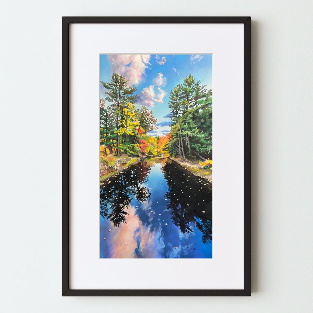 A mockup of a landscape art print in a black frame with a white mat. The painting is tall and narrow, showing a river scene with trees on either side. It's mostly blue water and sky, with autumn leaves, and looks like a very symmetrical piece.