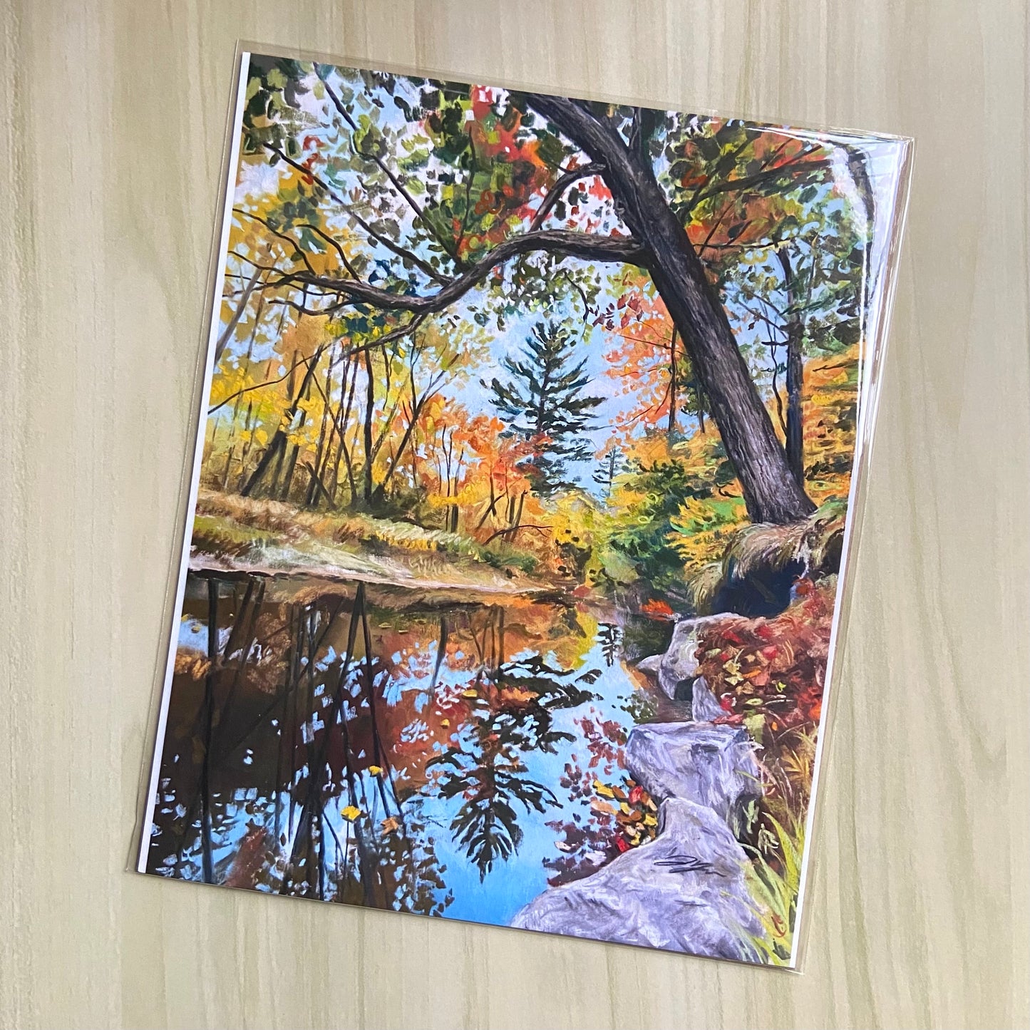 An art print of a painting that shows a calm river in a forest with autumn colors. A tree close by grows out over the river at an angle. The reflection of the trees in the water is darker than the real trees, creating depth in the river.