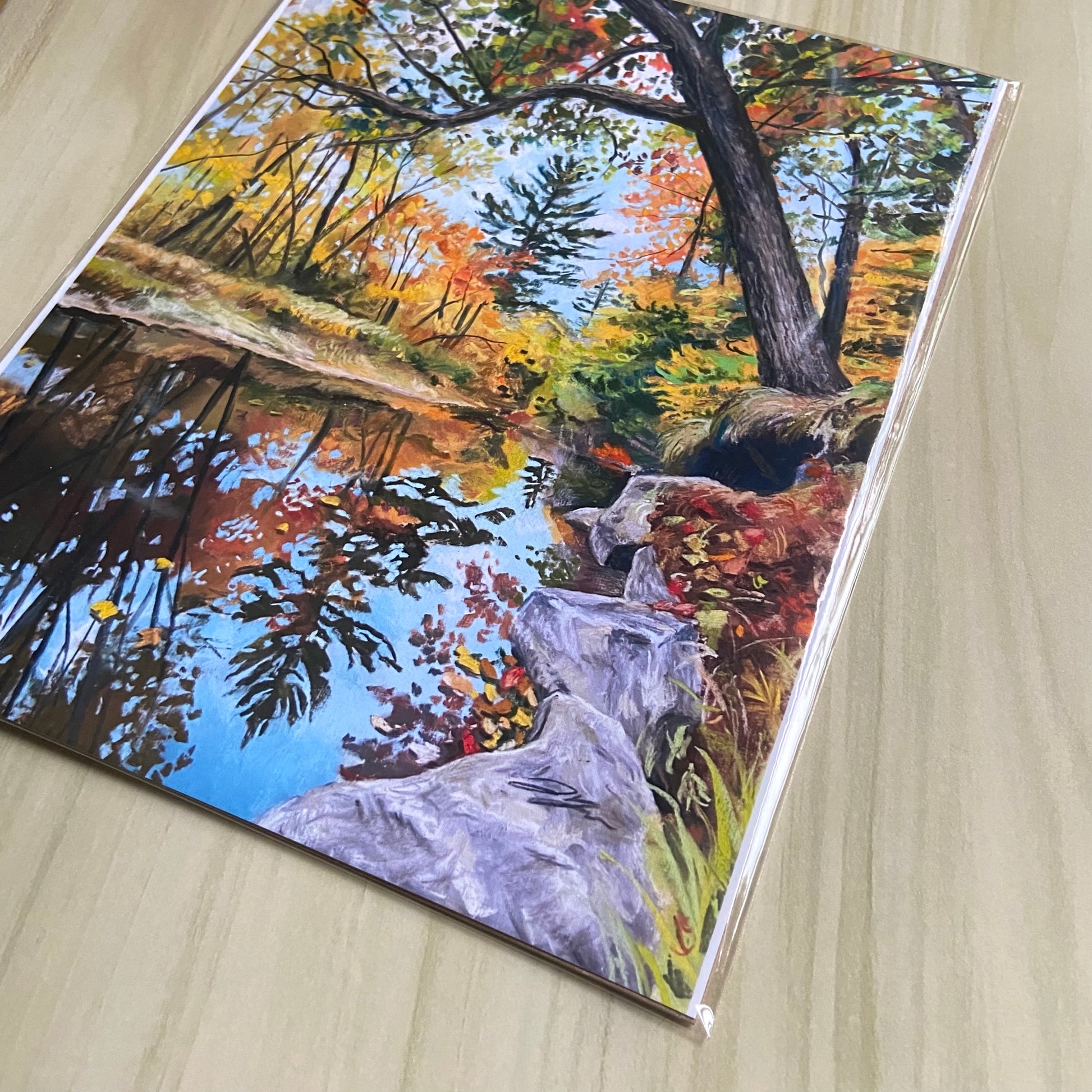 A detail of the landscape picture. The rocks by the water are close to the camera here, and the artist's signature can be seen. The painting is vibrant, with lots of sky blue and fall yellow, orange and red. The original is made with soft pastels.